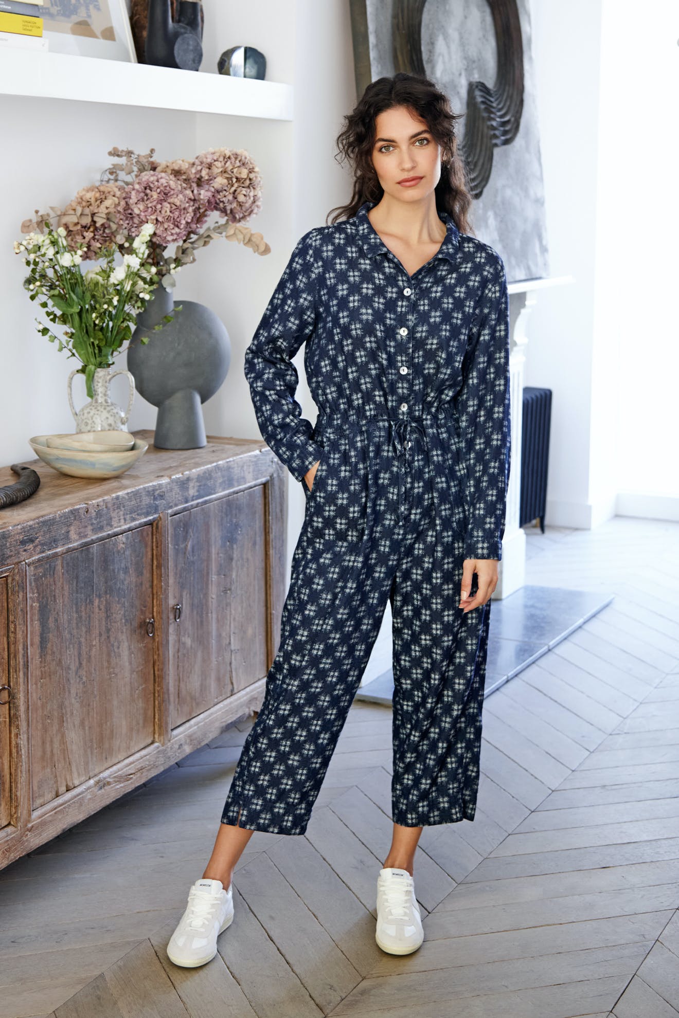 A woman stands in a room, wearing a Broken Check Denim Jumpsuit with an adjustable tie waist and white sneakers, with one hand on her hip and a slight smile.