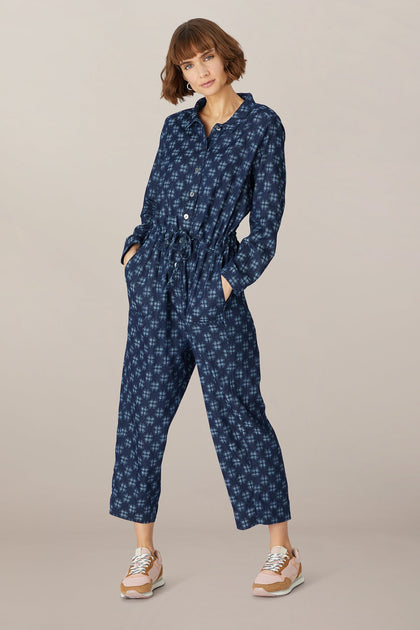 Woman in a Broken Check denim jumpsuit with pockets, standing against a neutral backdrop, hands in pockets, looking at the camera.