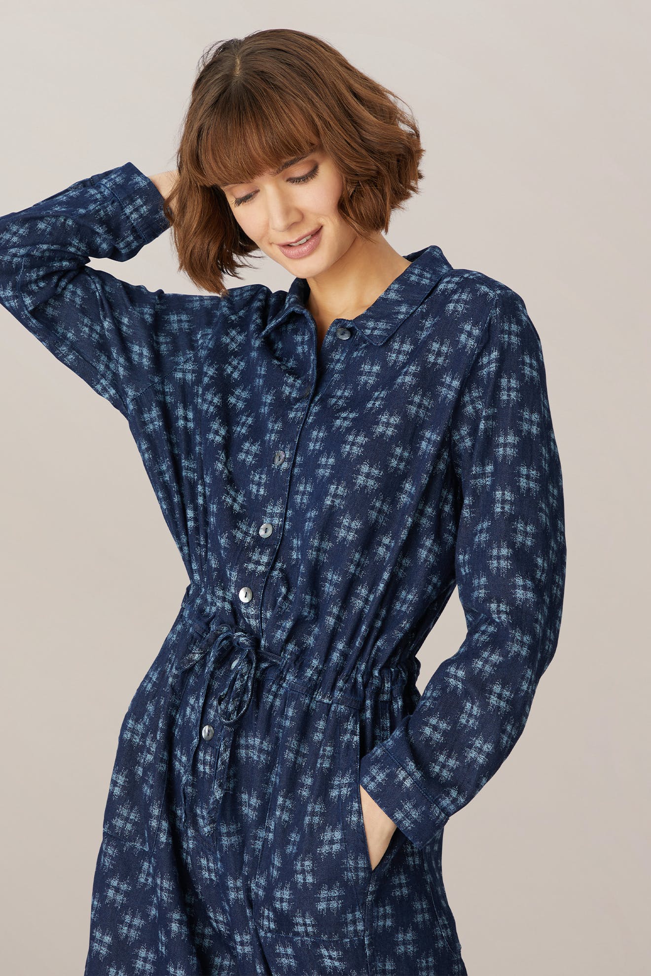 A woman with short brown hair, smiling gently, wearing a Broken Check denim jumpsuit with an adjustable tie waist, posing with her hand on her head against a neutral background.