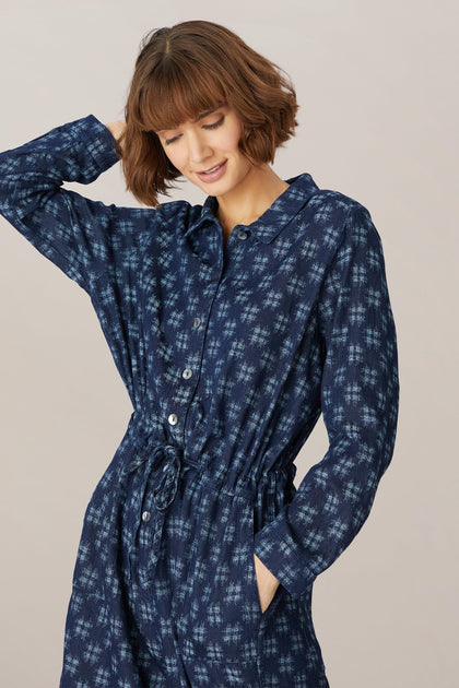 A woman with short brown hair, smiling gently, wearing a Broken Check denim jumpsuit with an adjustable tie waist, posing with her hand on her head against a neutral background.