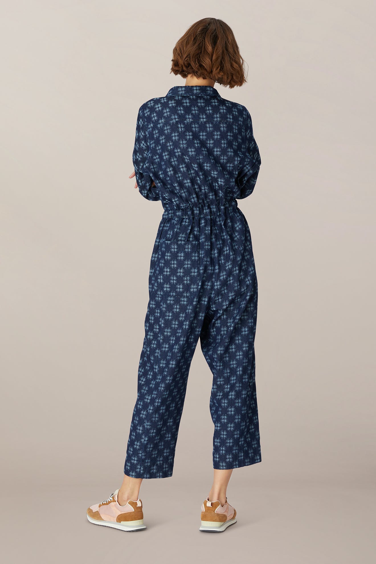 A woman stands with her back to the camera, wearing a Broken Check Denim Jumpsuit and white sneakers against a neutral background.