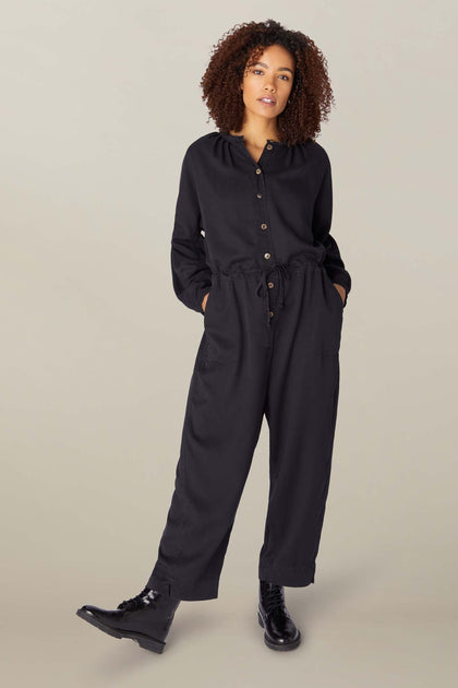 A woman in a Drape Tencel Twill jumpsuit and glossy boots stands on a beige background, looking at the camera.