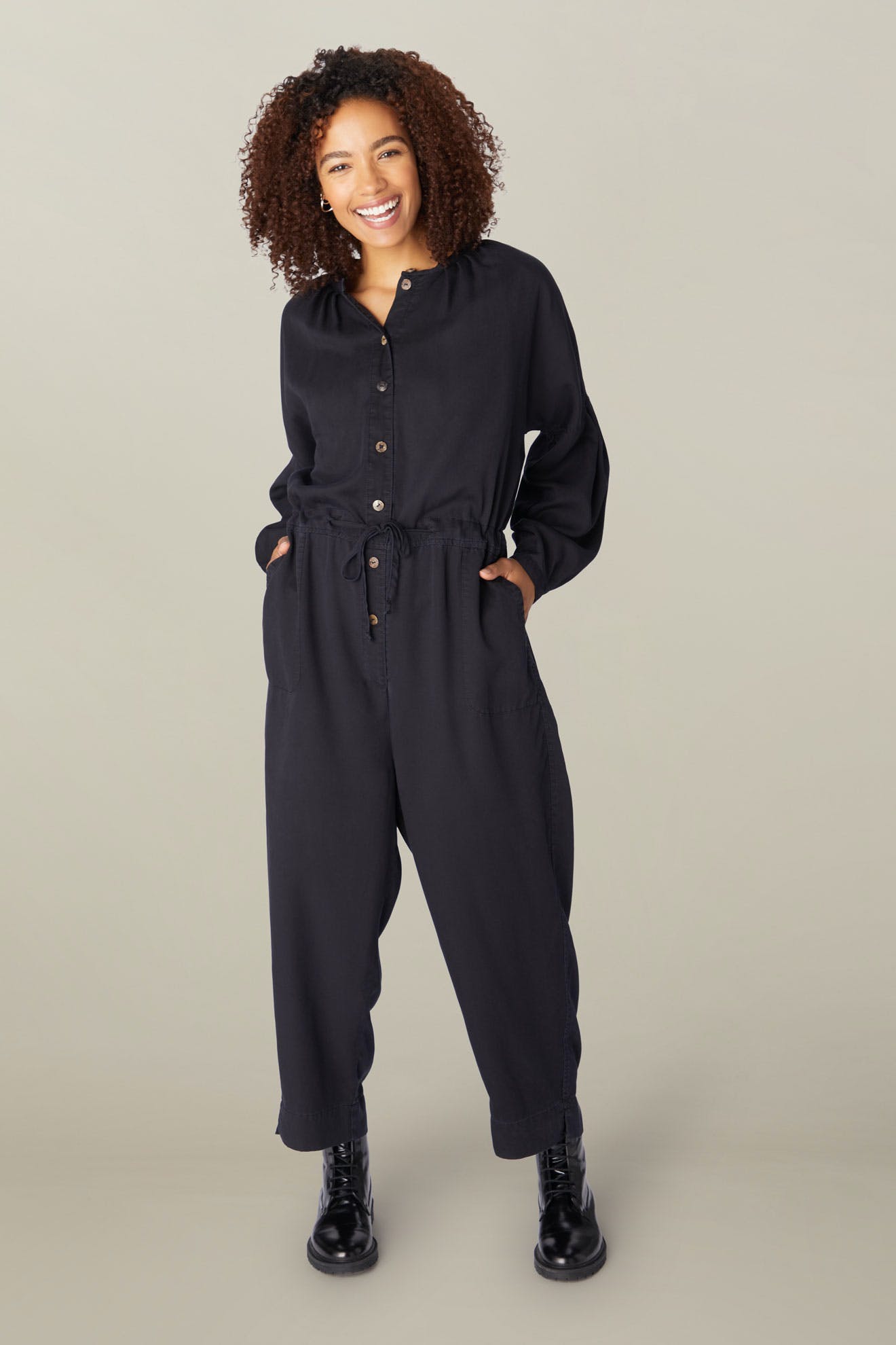 A woman with curly hair, smiling in a studio, wearing a dark Drape Tencel Twill Jumpsuit with pockets and black boots, standing with hands in pockets.