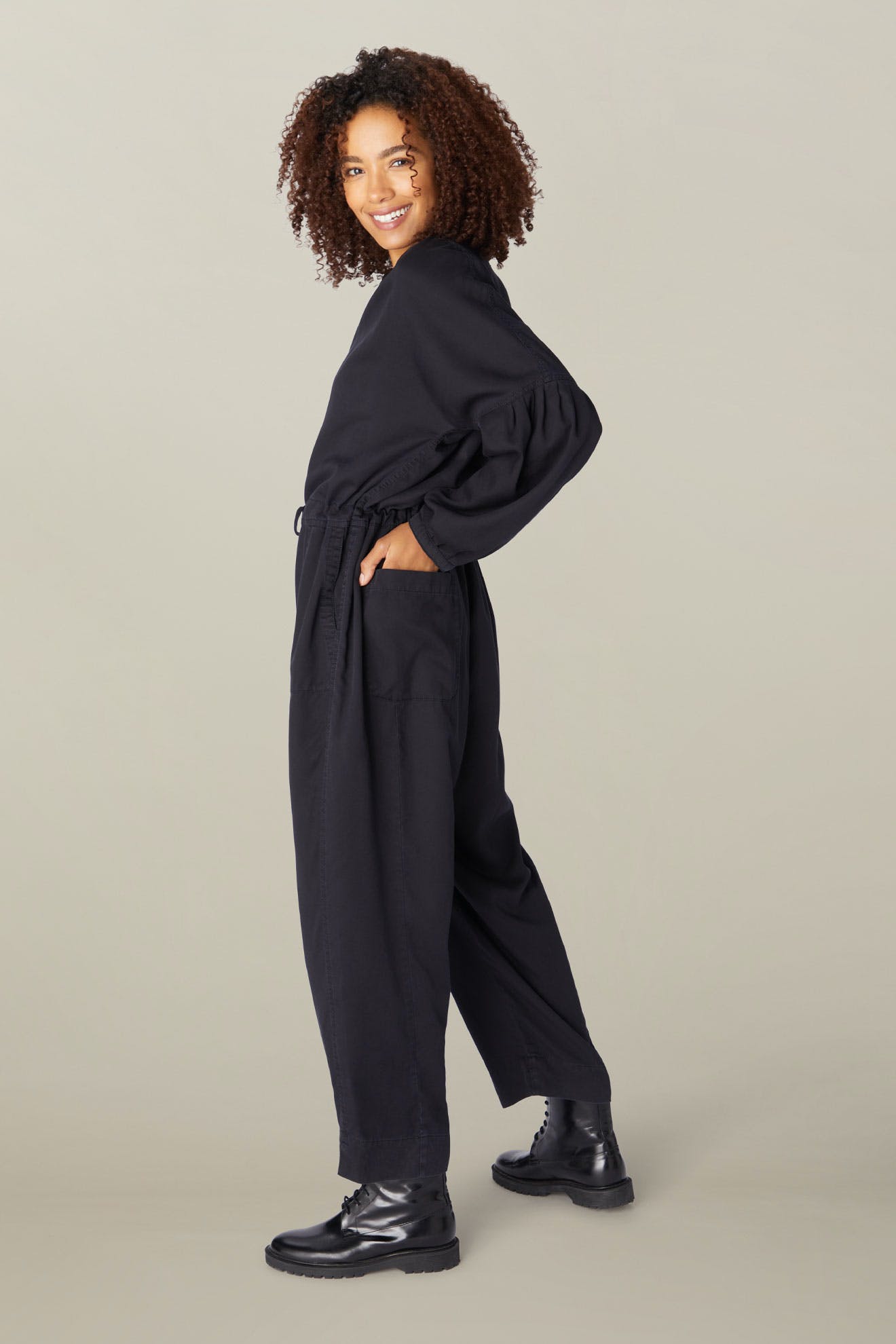 A woman with curly hair smiles, wearing a black Drape Tencel Twill Jumpsuit and boots, standing side-on with hands in pockets against a beige background.
