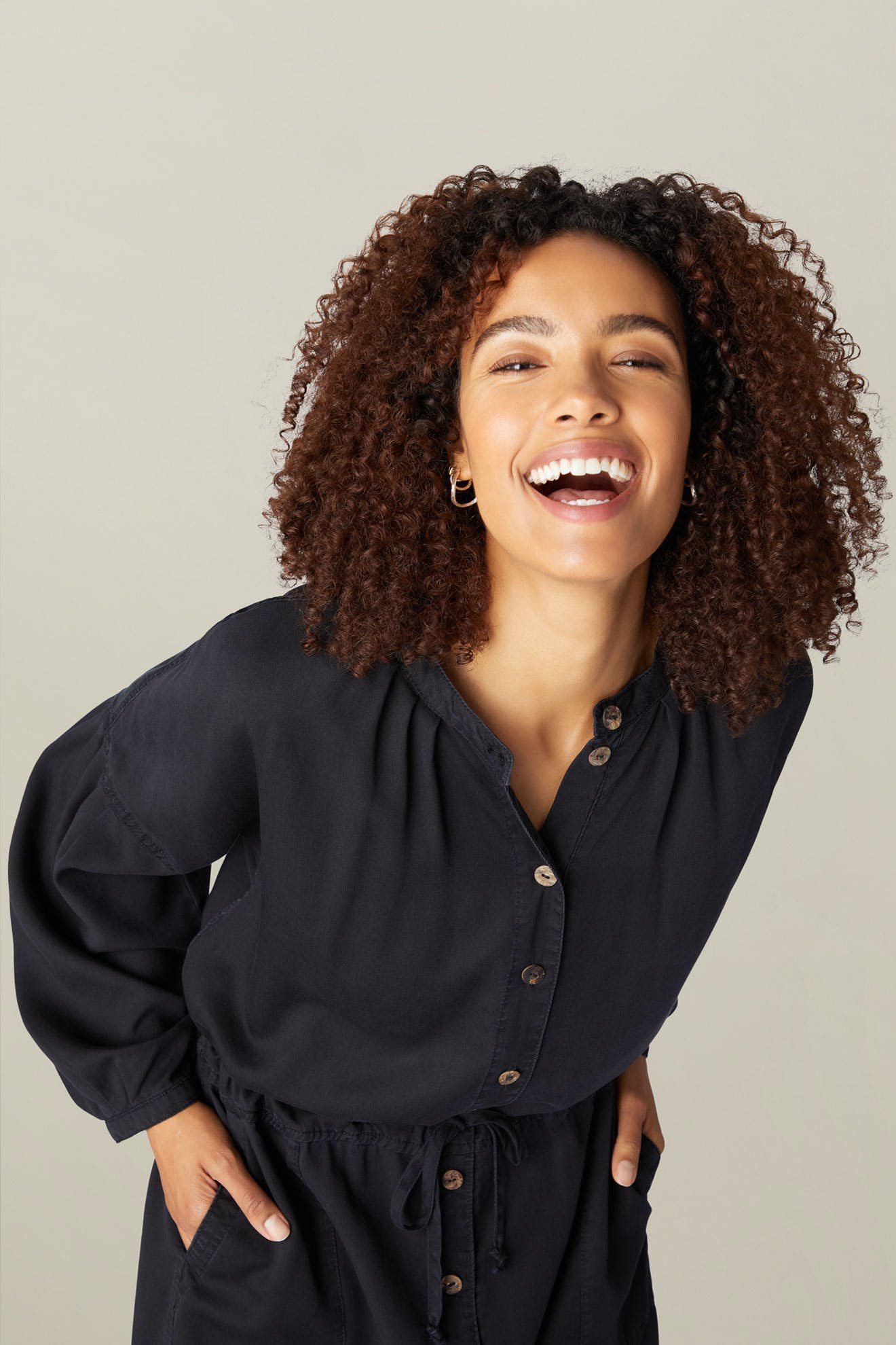 A joyful woman with curly hair, wearing a Drape Tencel Twill Jumpsuit, laughing while posing with one hand on her hip against a light gray background.