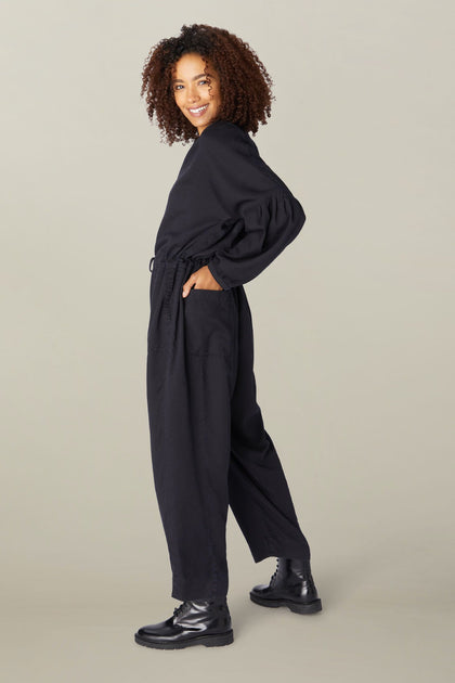 A woman with curly hair smiling at the camera, wearing a Drape Tencel Twill Jumpsuit and black boots, standing sideways with one hand on her hip.