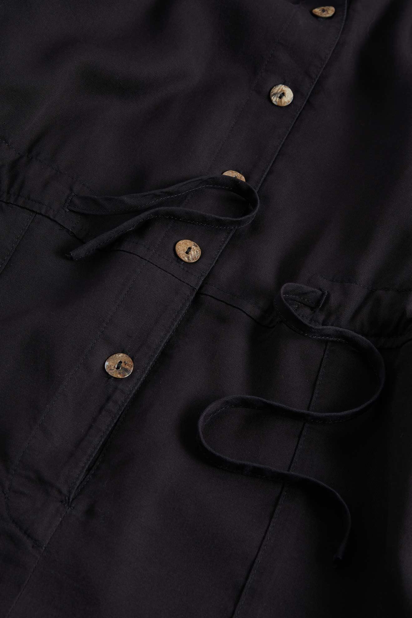 Close-up of a black trench coat focusing on button details and a Drape Tencel Twill Jumpsuit.