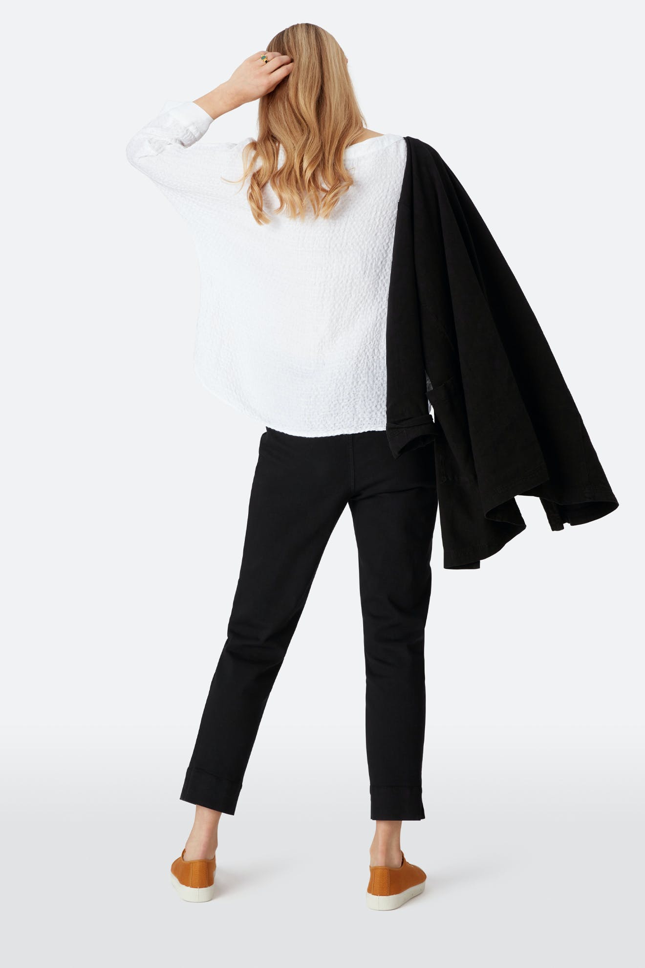 A person stands with their back to the camera, wearing a white textured shirt, tan shoes, holding a black coat over one shoulder, and black Stretch Cotton Twill Trousers with an elasticated waist and slim tapered legs.