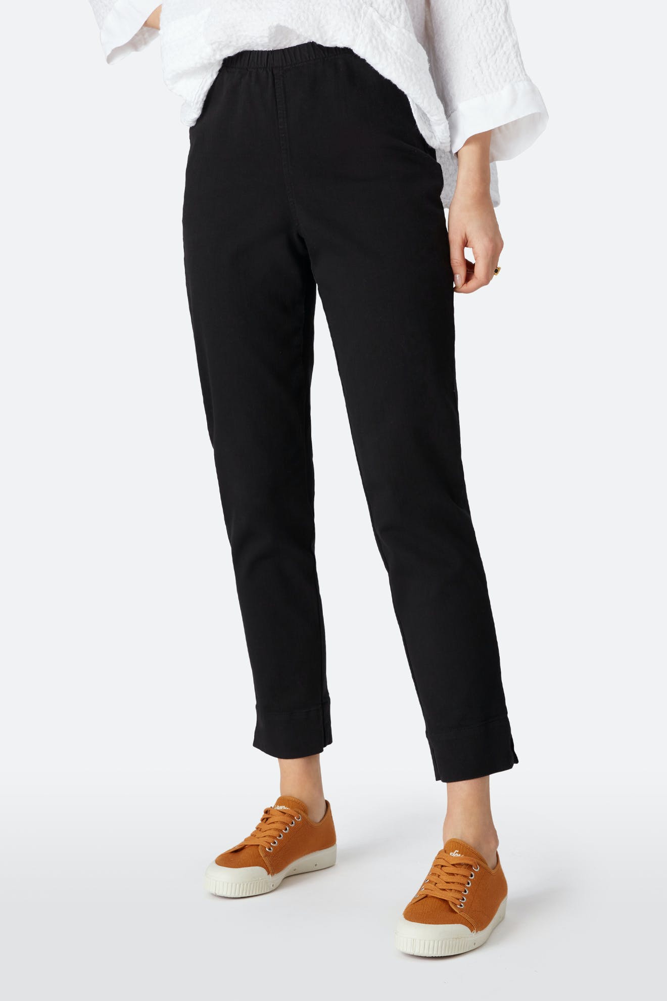 Person wearing black Stretch Cotton Twill Trouser with an elasticated waist, a loose white top, and brown sneakers, standing against a plain light background.