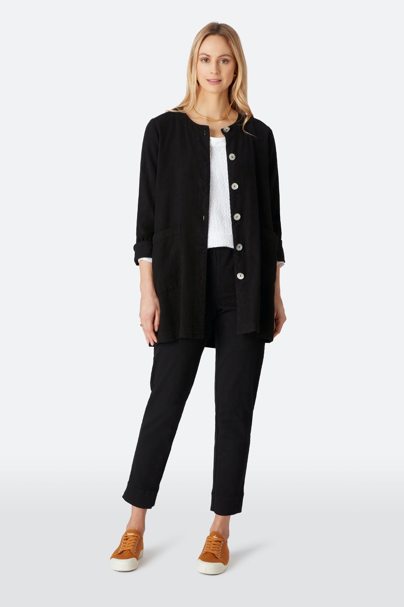 A woman faces forward in a black button-up long jacket, a white top, mustard yellow sneakers, and Stretch Cotton Twill Trousers with a slim-leg tapered fit and elasticated waist against a plain light background.