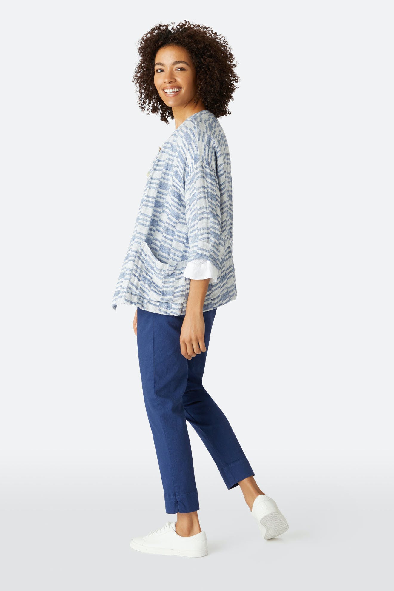 Person standing and smiling, wearing a loose blue-and-white patterned top, Stretch Cotton Twill Trouser with an elasticated waist and slim-leg tapered fit, plus white sneakers, against a plain light background.