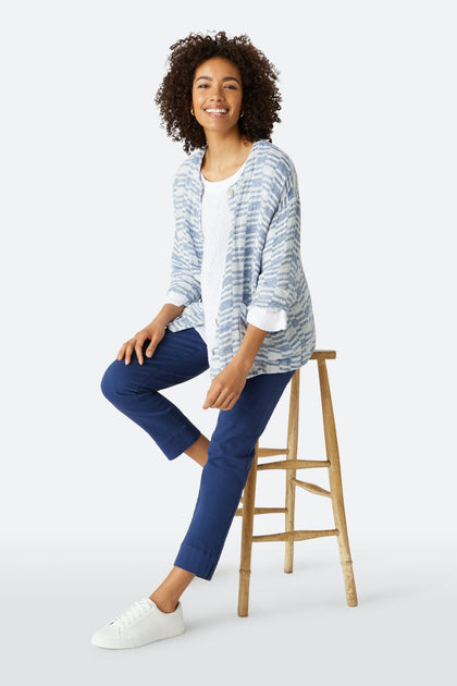 A woman sits on a wooden stool in a white shirt, blue and white striped cardigan, Stretch Cotton Twill Trouser with an elasticated waist, and white sneakers, smiling at the camera against a plain background.