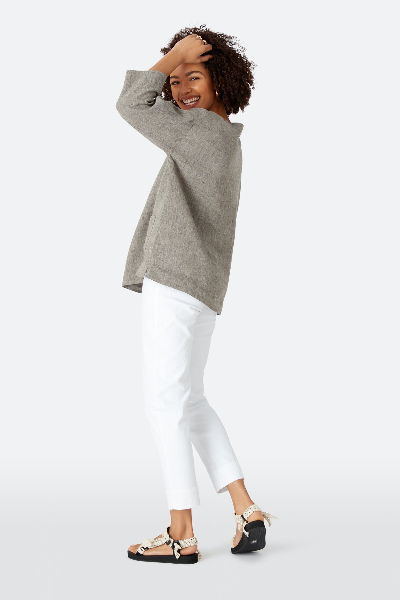 A woman stands sideways, smiling and touching her hair. She wears a loose gray top, black sandals with rope detailing, and Stretch Cotton Twill Trousers with a slim-leg tapered fit and elasticated waist against a plain light background.