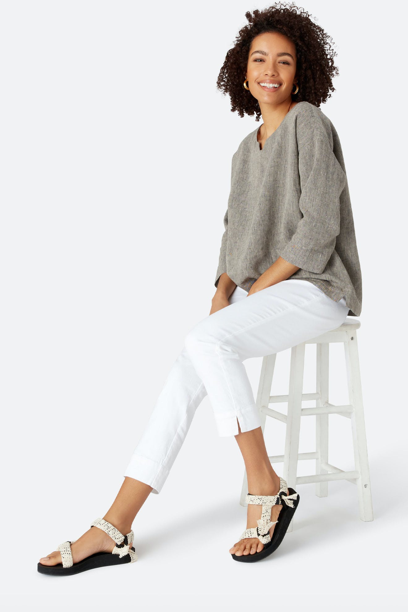 A woman sits on a white stool, smiling at the camera in a gray long-sleeve top and Stretch Cotton Twill Trouser with an elasticated waist, paired with beige sandals with black soles.