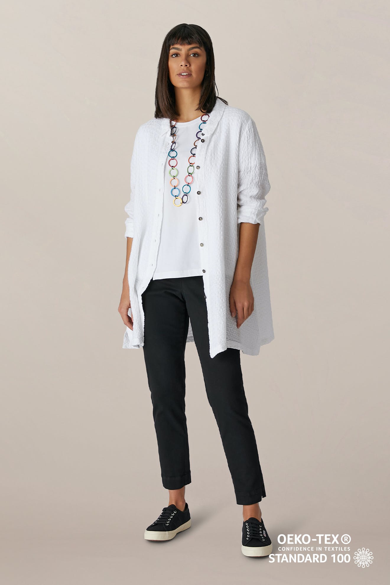 A woman wears a long white button-up shirt over a white top, SS22 Stretch Cotton Trousers with an elasticated waist, black sneakers, and a colorful necklace. The OEKO-TEX STANDARD 100 logo appears in the corner.