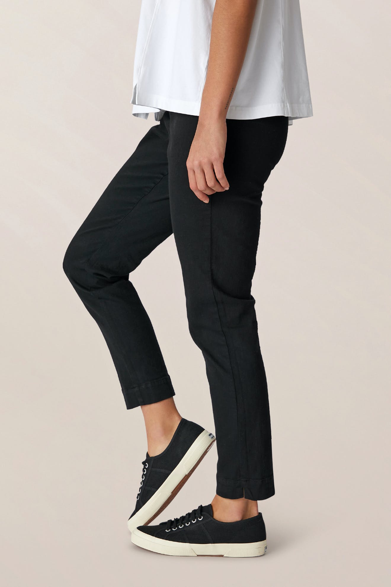 A person is wearing the SS22 Stretch Cotton Trouser—black, slim-leg with an elasticated waist—paired with a white top and black sneakers with white soles, standing against a plain, light-colored background.