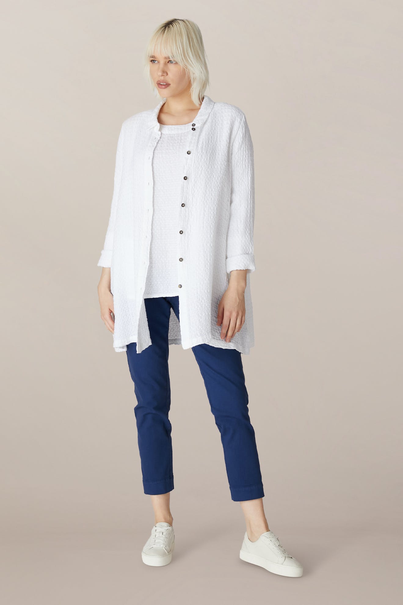 A person stands facing forward, wearing a white long button-up shirt over a white top, SS22 Stretch Cotton Trousers in blue with an elasticated waist, and white sneakers against a plain background.
