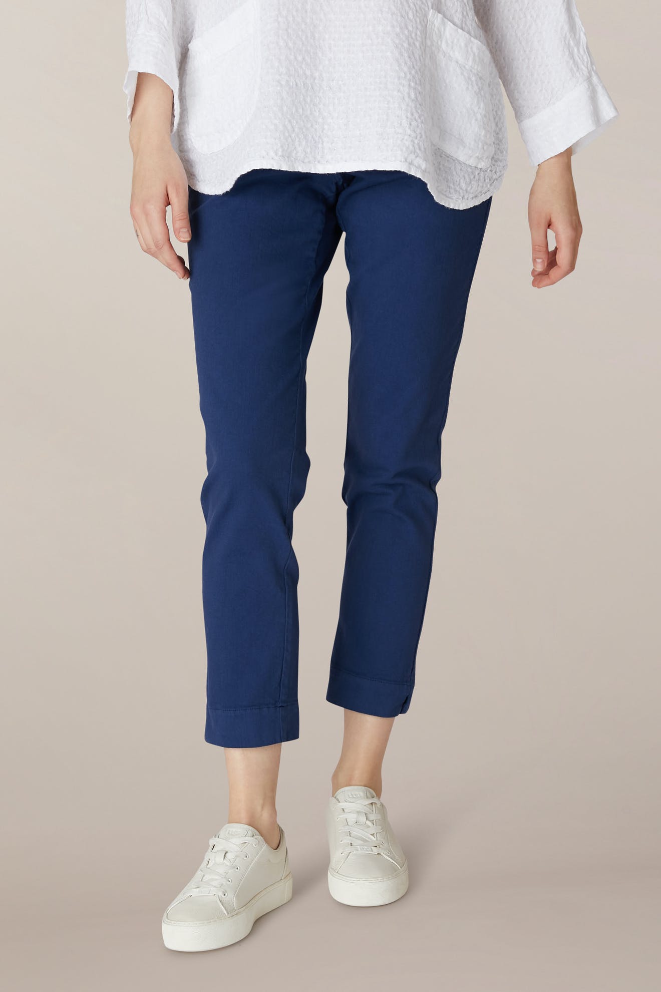 A person stands against a plain background wearing a white textured long-sleeve top, SS22 Stretch Cotton Trouser in blue with an elasticated waist, and white sneakers.