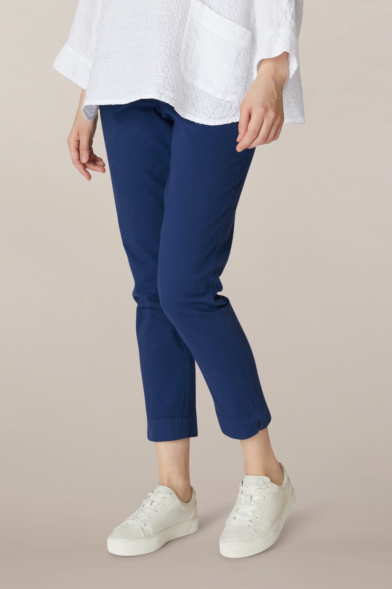 Someone stands against a light beige background, wearing a white textured top, white sneakers, and the SS22 Stretch Cotton Trouser—slim-leg and tapered with an elasticated waist.