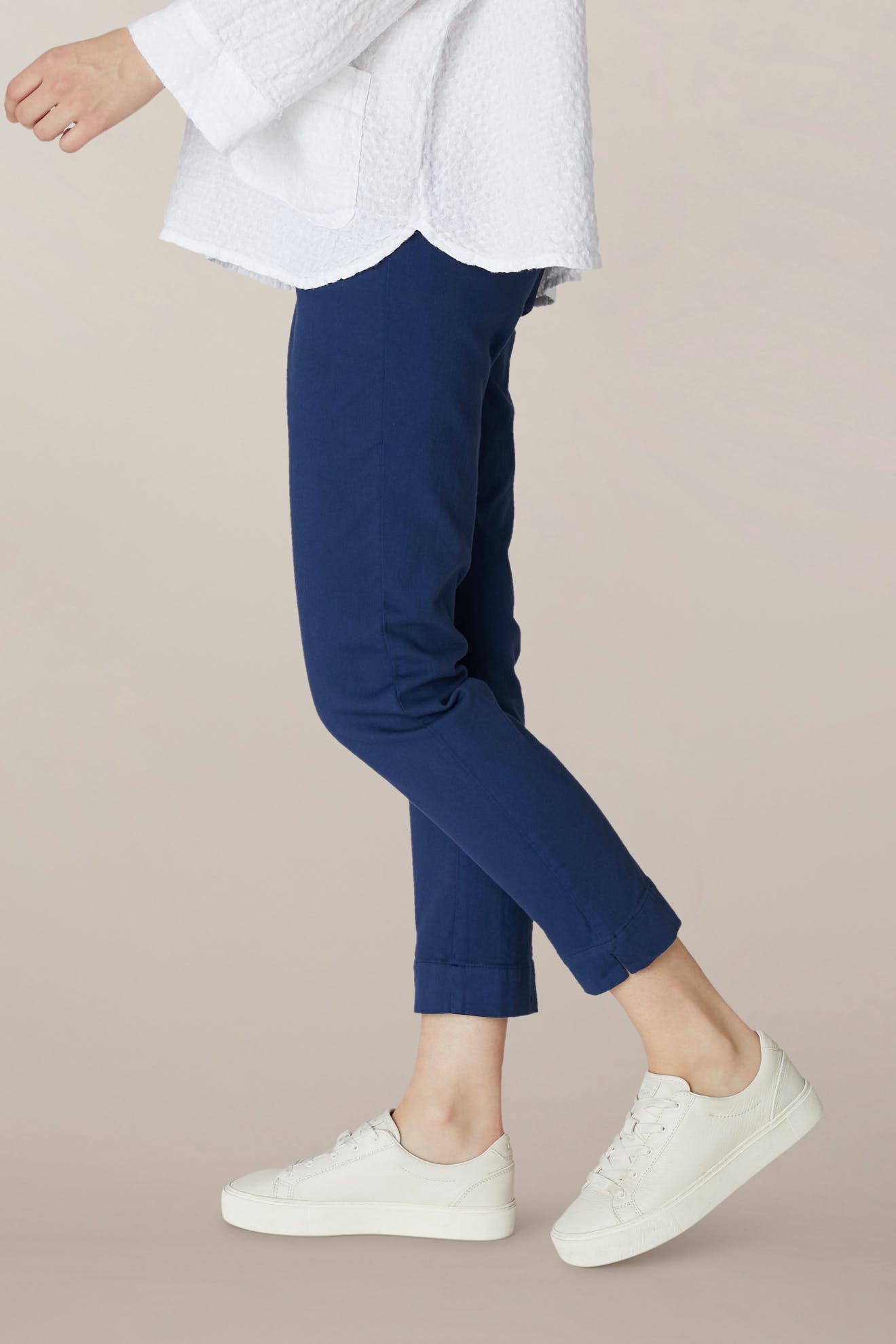 Wearing SS22 Stretch Cotton Trouser in blue with an elasticated waist, a white textured long-sleeve top, and white sneakers, standing against a plain beige background.
