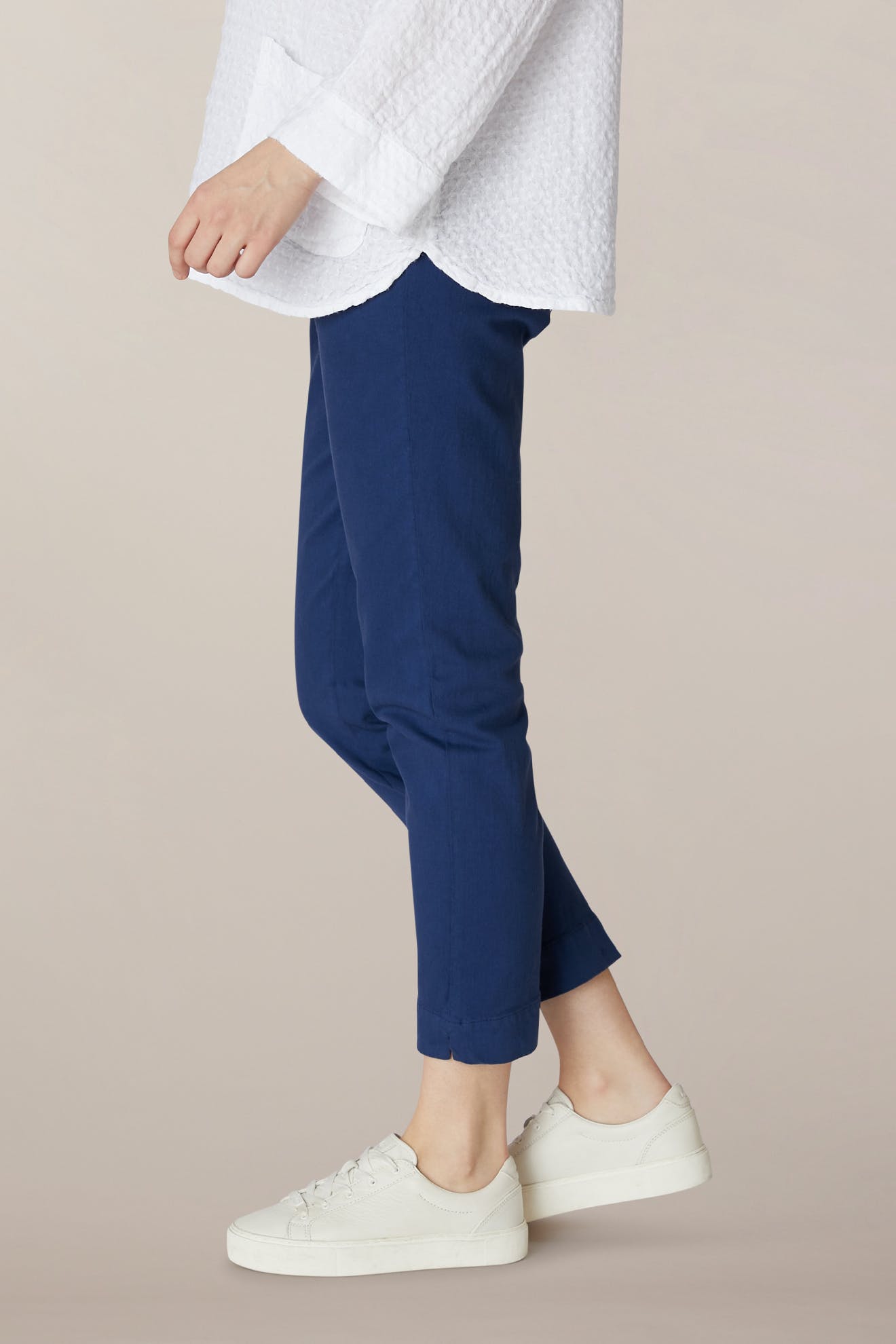 Person wearing a white textured long-sleeve top, SS22 Stretch Cotton Trouser in blue with an elasticated waist, and white sneakers, standing against a plain light background.