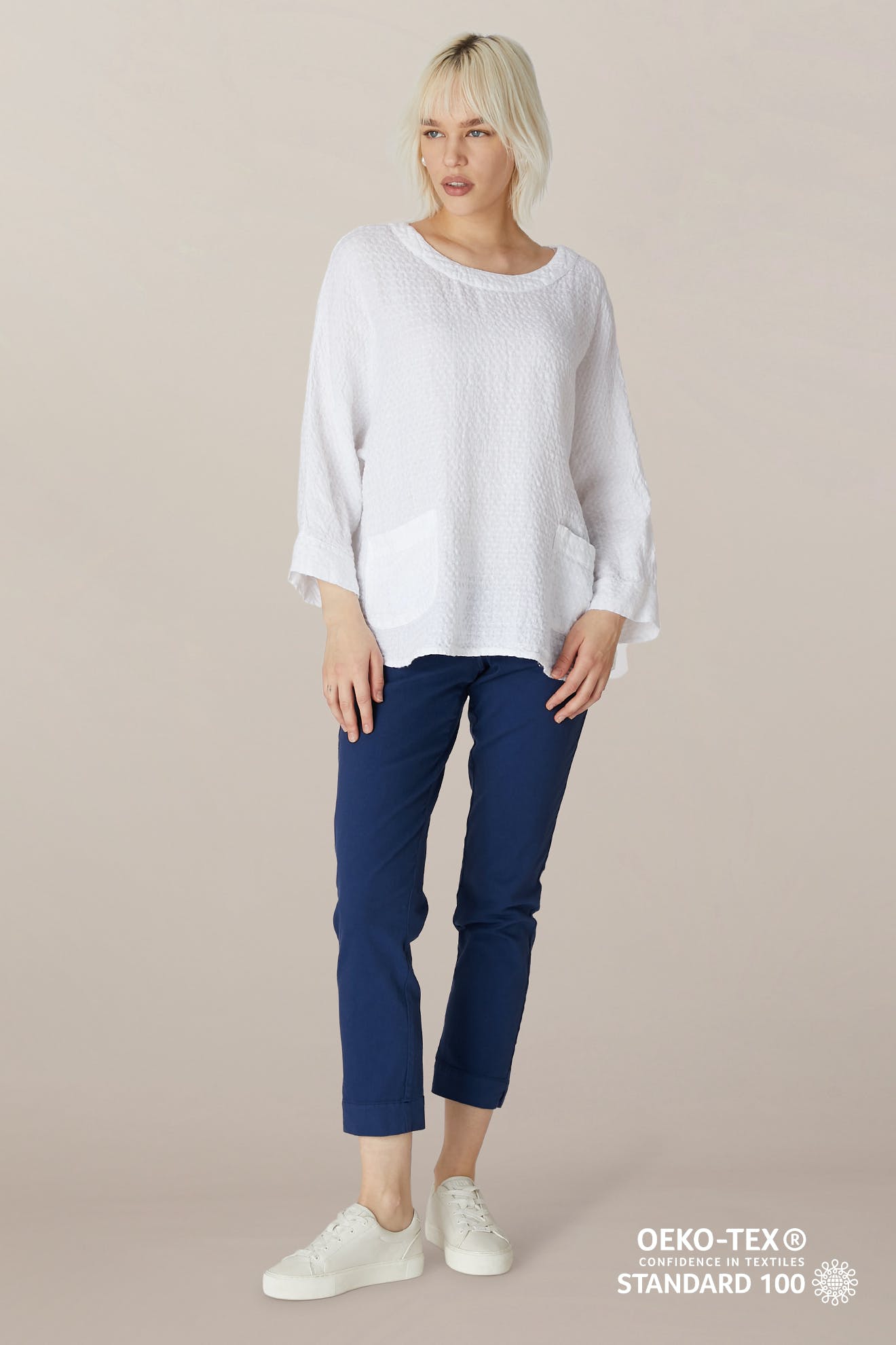 Woman wears a loose white textured long-sleeve top with front pockets, SS22 Stretch Cotton Trouser in blue (OEKO-TEX STANDARD 100), featuring slim tapered legs and elasticated waist, and white sneakers, against a neutral background.