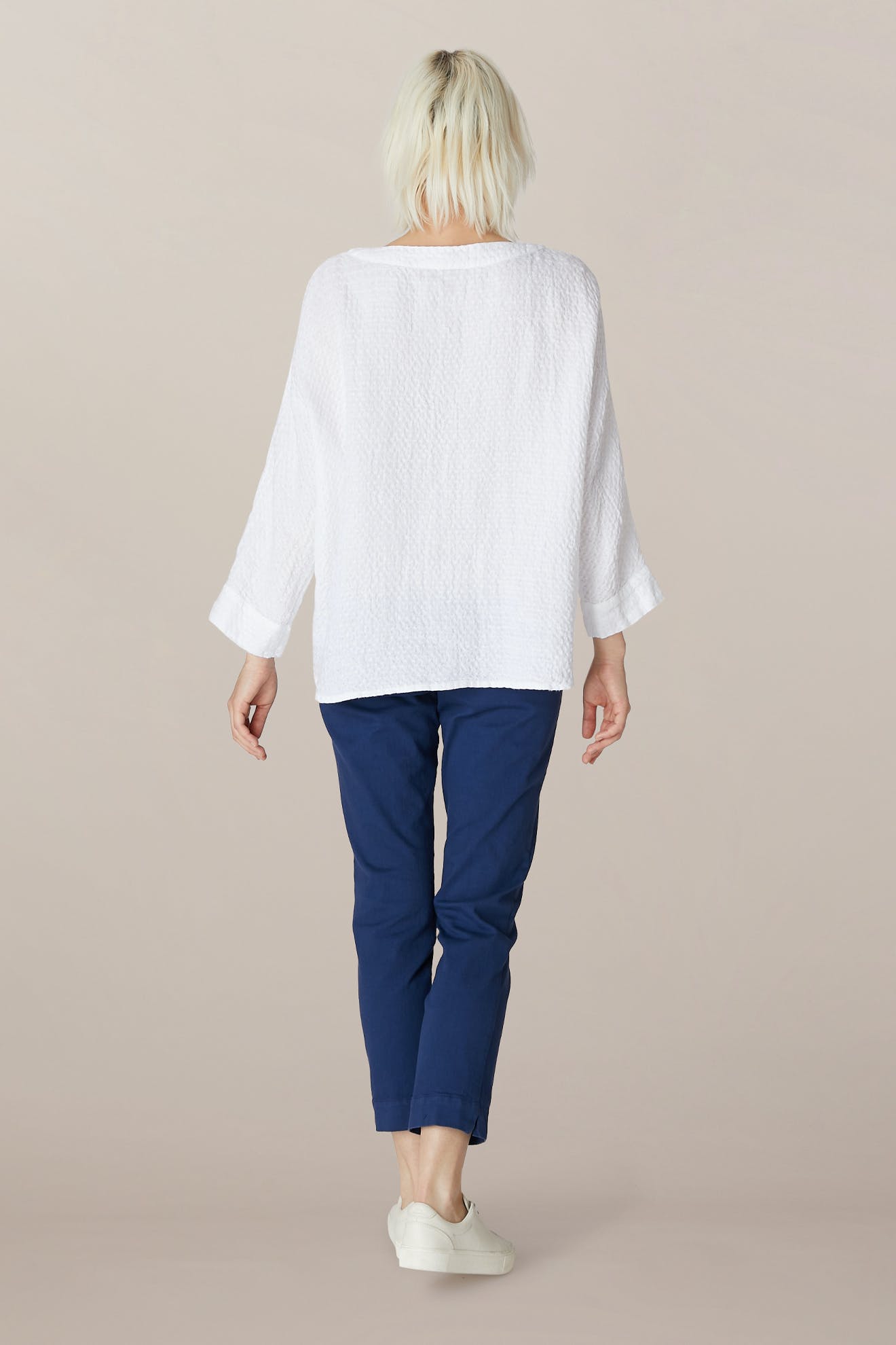 A person with short blonde hair, wearing a loose white long-sleeve top, SS22 Stretch Cotton Trouser with an elasticated waist and slim tapered fit, and white shoes, stands facing away against a plain background.
