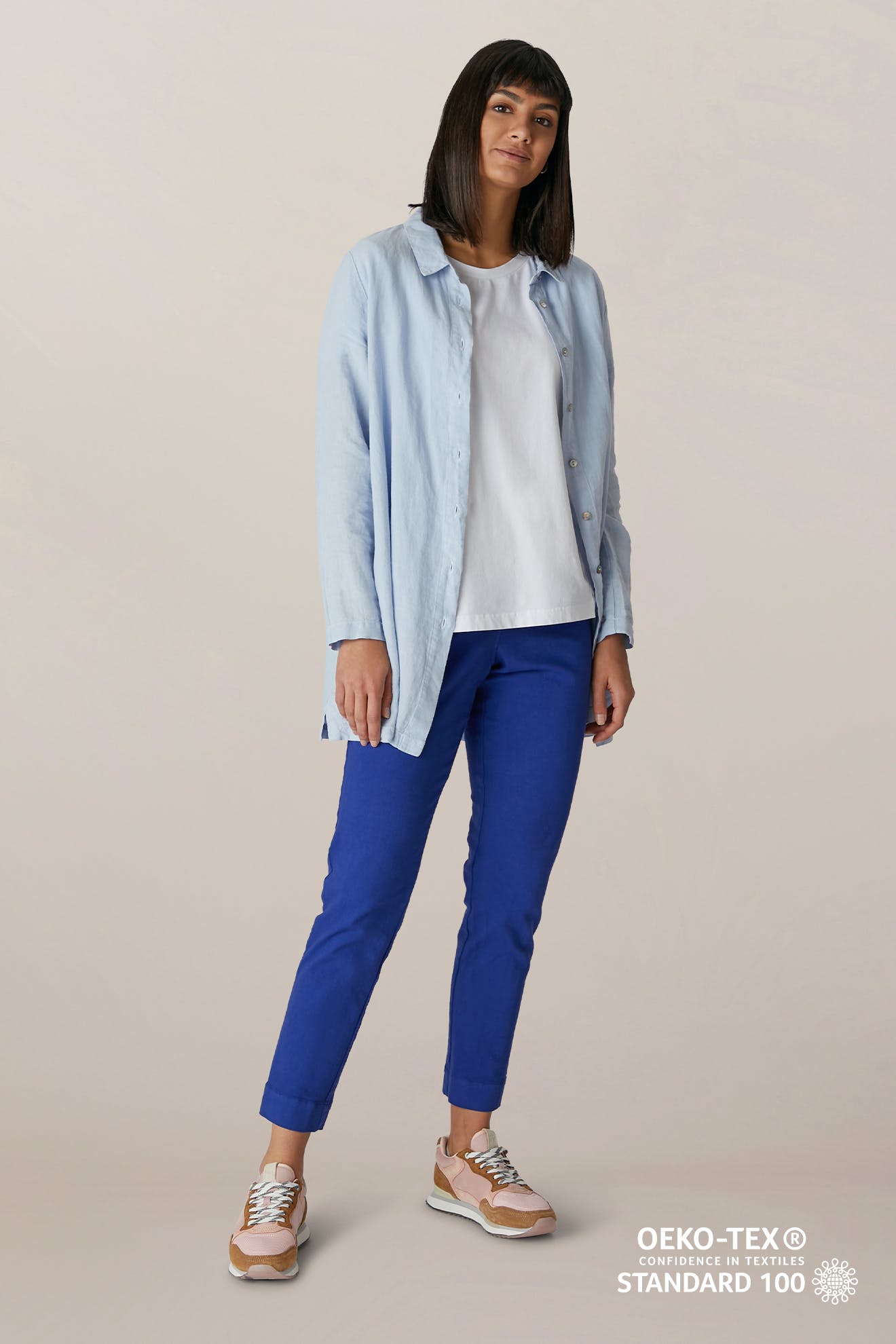 A woman stands against a neutral background wearing a light blue button-up shirt, white tee, and the SS22 Stretch Cotton Trouser with an elasticated waist. Pink sneakers complete the look. The OEKO-TEX Standard 100 logo is shown at the bottom.