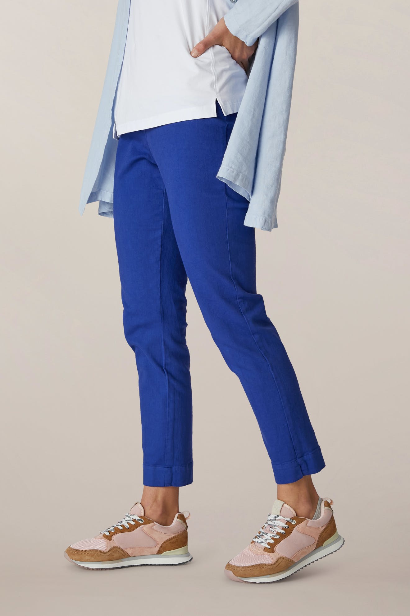 Person wearing SS22 Stretch Cotton Trouser in blue, a white OEKO-TEX cotton shirt, light blue overshirt, and pink sneakers, standing with one hand in pocket against a plain background.