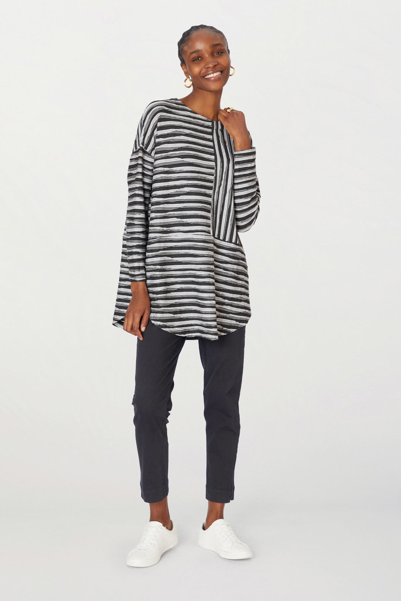 A smiling woman stands in a studio wearing a striped tunic, Stretch Twill Cotton Trousers, and white sneakers.