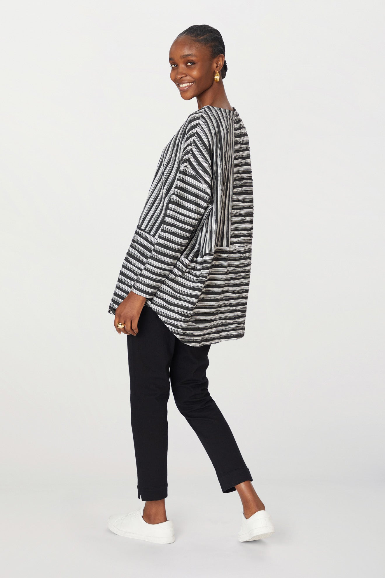 A woman in a striped cardigan, stretch twill cotton trousers, and white sneakers walks right, smiling, looking over her shoulder.
