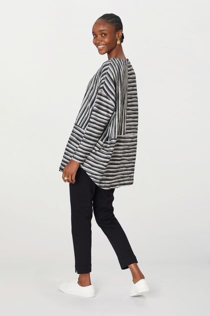 A woman in a striped cardigan, stretch twill cotton trousers, and white sneakers walks right, smiling, looking over her shoulder.