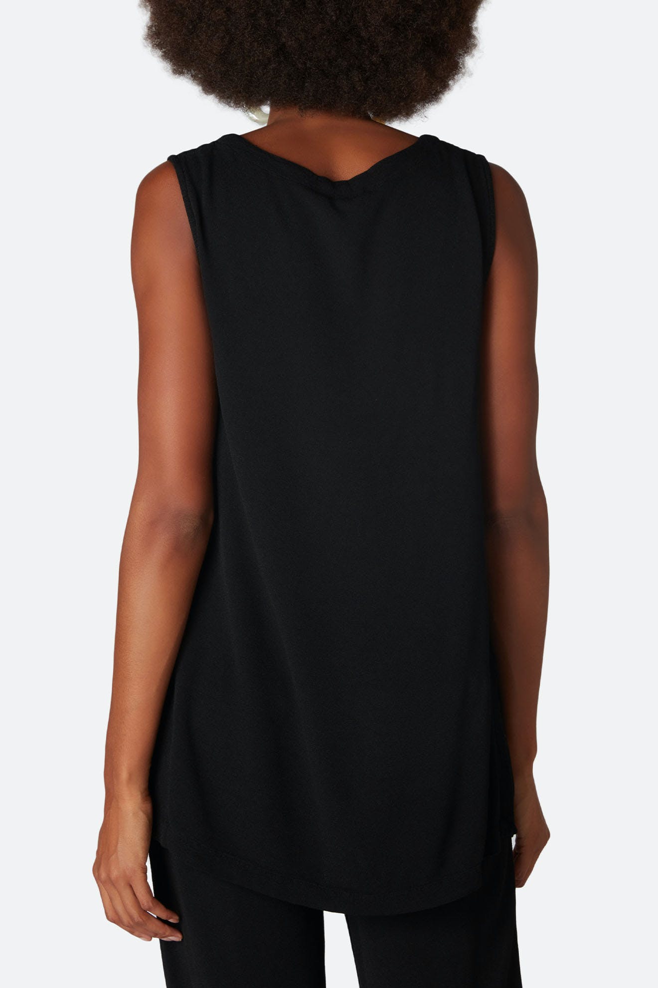 A person with curly hair, viewed from the back, is wearing a Crepe Long Vest in black paired with black pants. The luxurious fabric creates a sleek longline silhouette against a plain background.