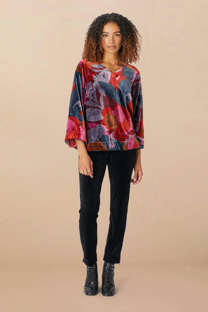 A person with curly hair wears the vibrant Winter Garden Velvet Top, featuring a scooped V neckline and a colorful floral design, paired with black pants and boots, standing against a beige background.