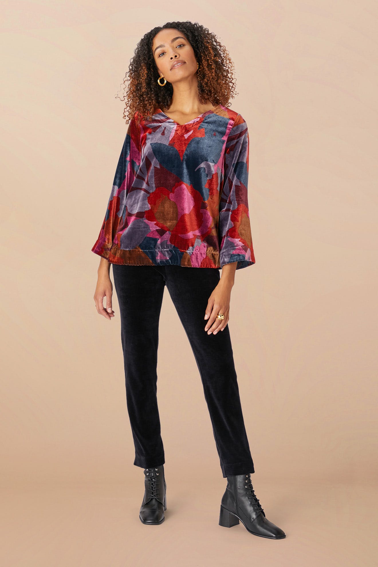 A person stands confidently, adorned in the Winter Garden Velvet Top, a floral blouse featuring long sleeves and a scooped V neckline, paired with black pants and black lace-up boots, against a neutral beige background.