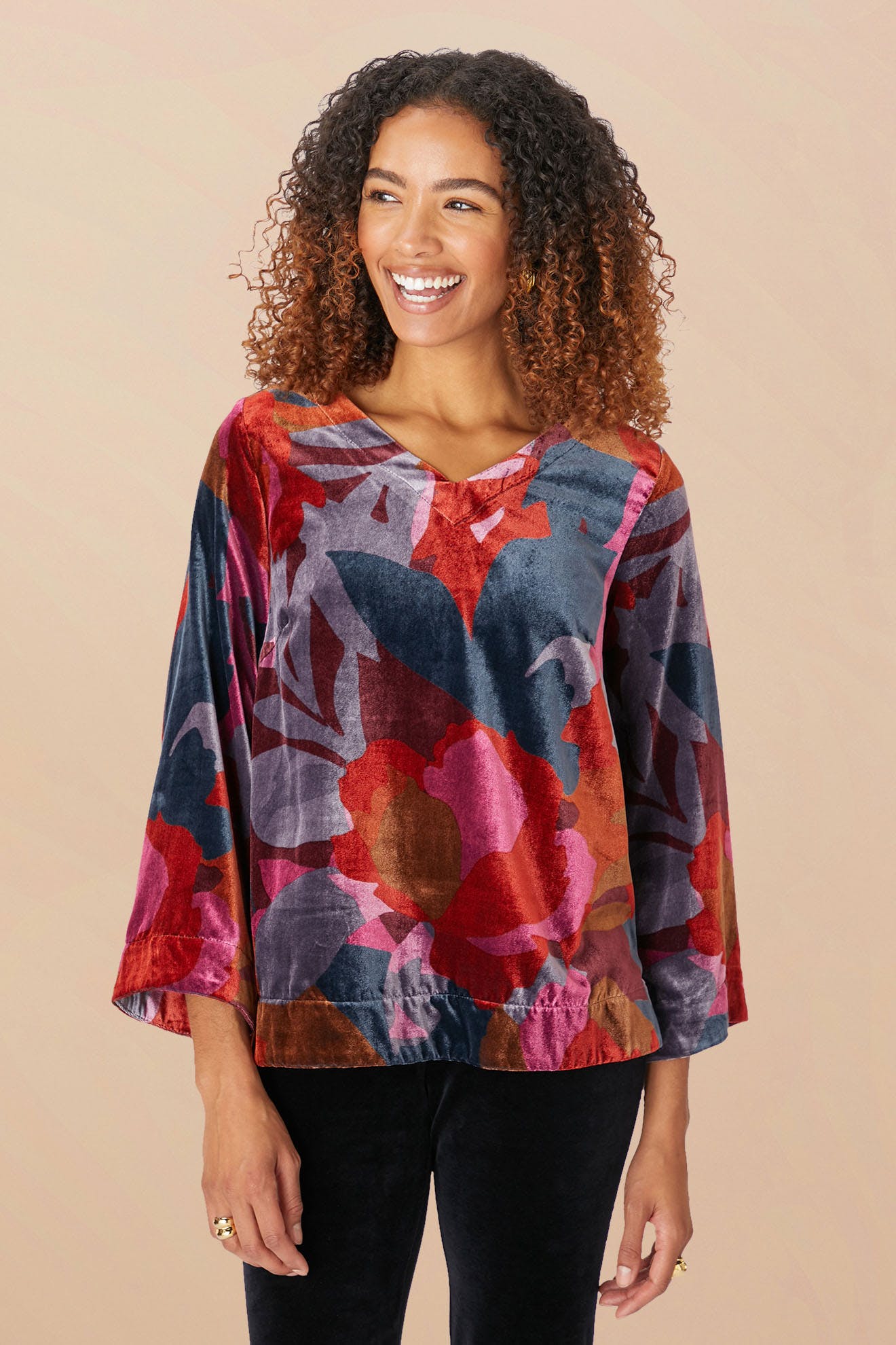 A person stands smiling, wearing the Winter Garden Velvet Top, a colorful floral-patterned long-sleeve blouse made of decadent velvet with a scooped V neckline, paired with black pants.