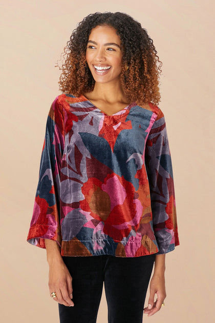 A person stands smiling, wearing the Winter Garden Velvet Top, a colorful floral-patterned long-sleeve blouse made of decadent velvet with a scooped V neckline, paired with black pants.