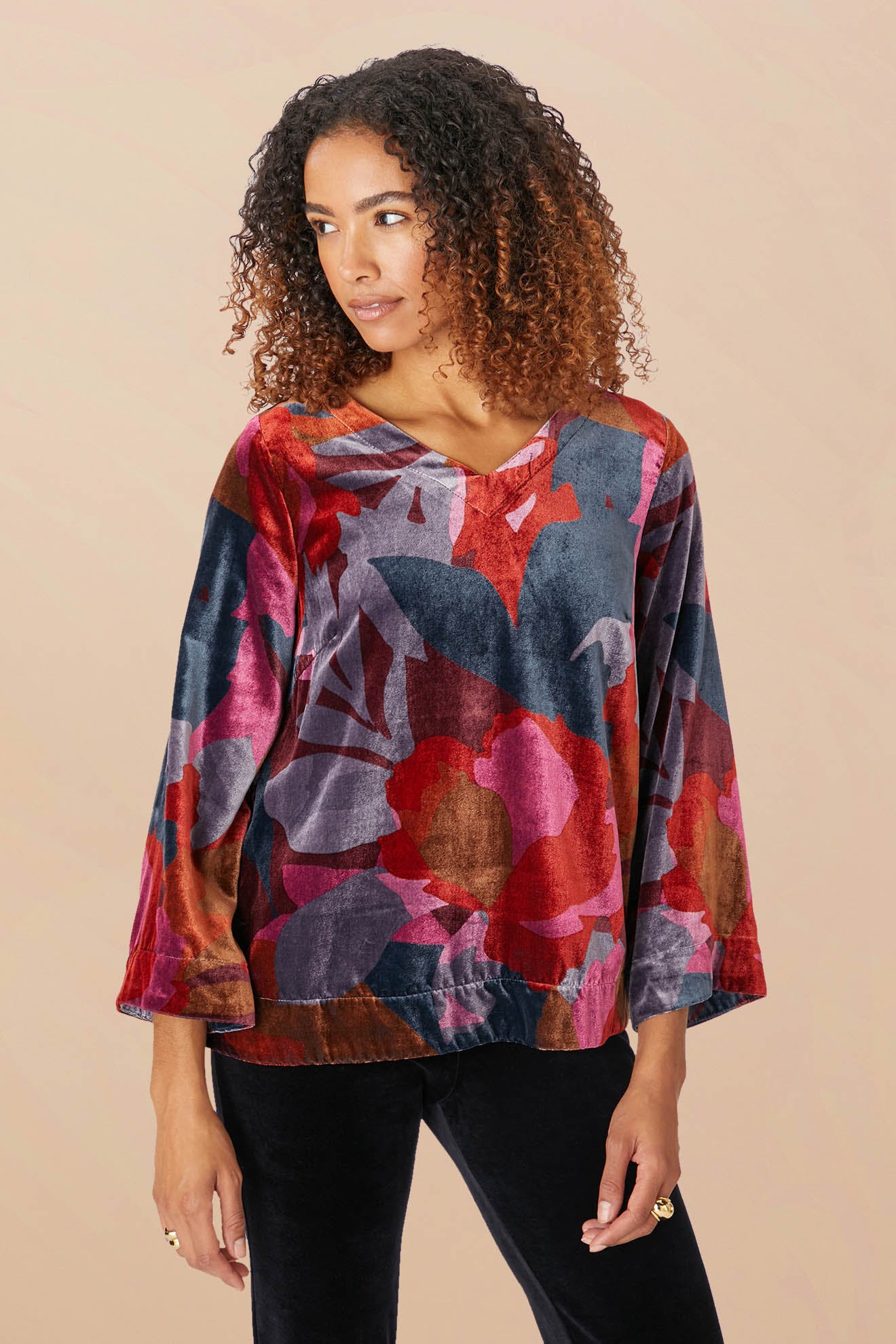 A person with curly hair wears the Winter Garden Velvet Top, a long-sleeved piece featuring a colorful abstract floral pattern and a scooped V neckline, paired with black pants, standing against a plain background.