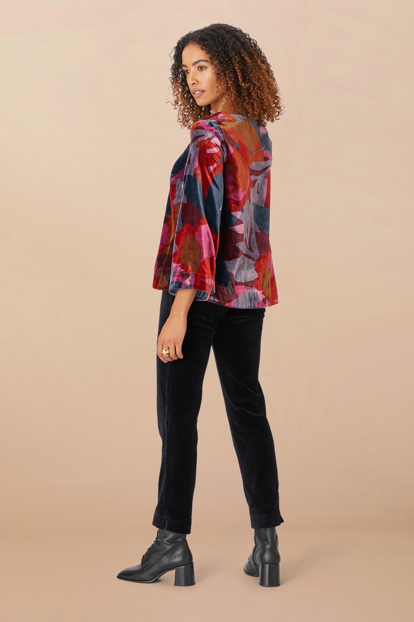 A person with curly hair stands sideways wearing the Winter Garden Velvet Top, featuring a floral print and scooped V neckline, paired with black pants and black heeled boots against a beige background.