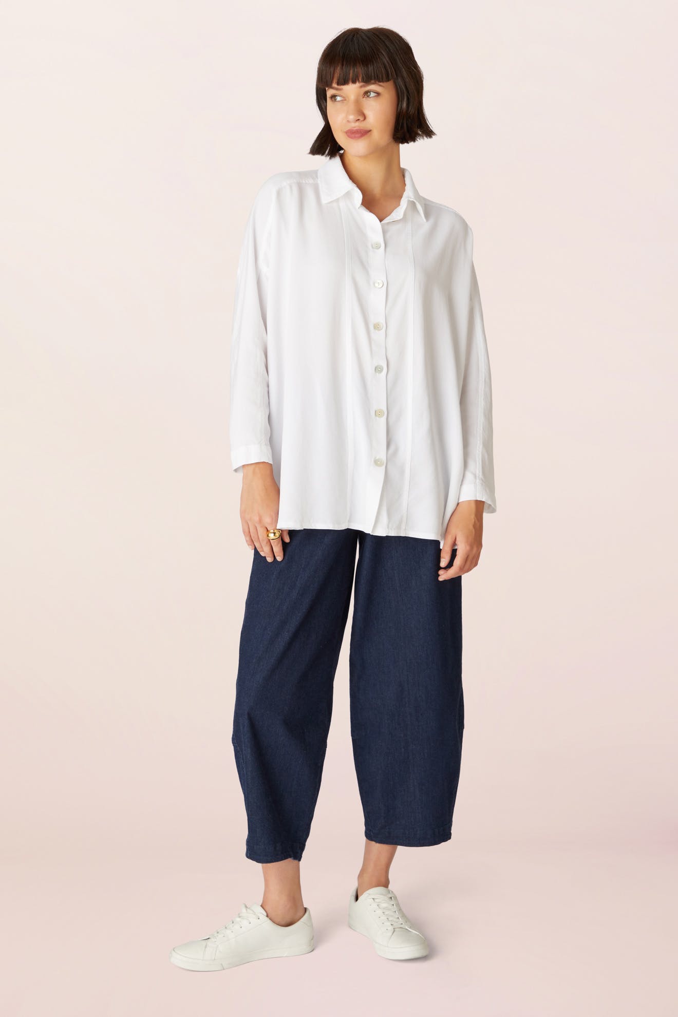 A woman stands wearing a loose, boxy Tencel Twill Boxy Shirt, wide-leg dark blue jeans, and white sneakers against a light background.