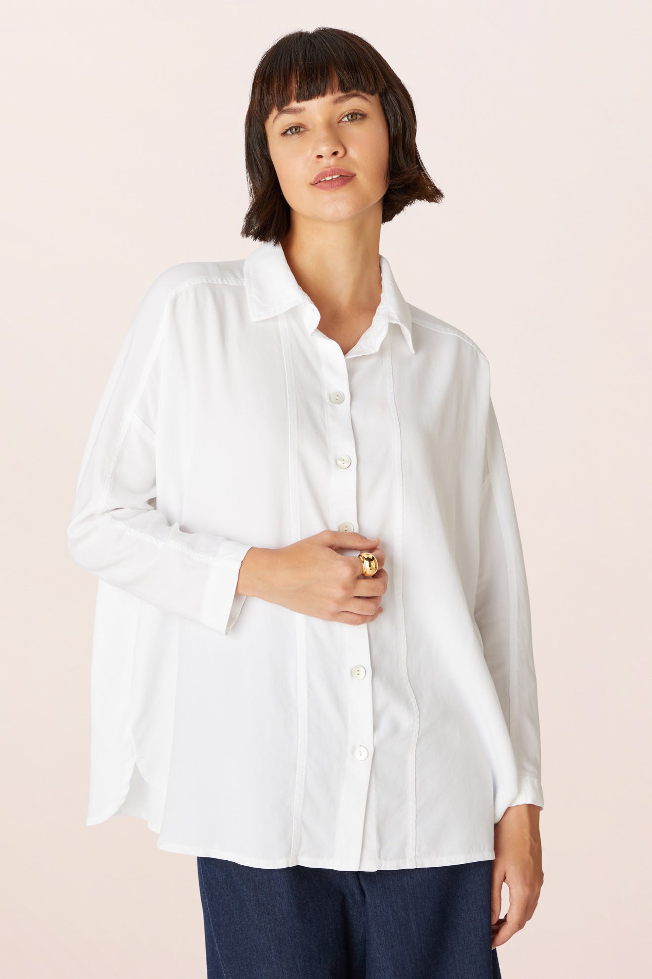 A person stands against a plain light background wearing the Tencel Twill Boxy Shirt—a loose-fitting, white sustainable long-sleeve top—paired with dark pants.