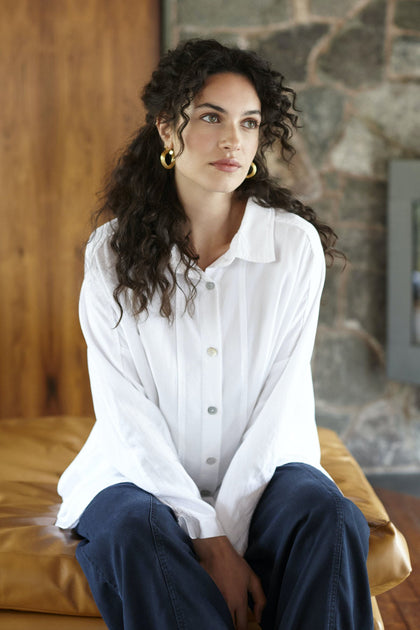 A woman with curly dark hair, wearing the Tencel Twill Boxy Shirt and blue jeans, sits on a tan couch in front of a stone and wood wall.