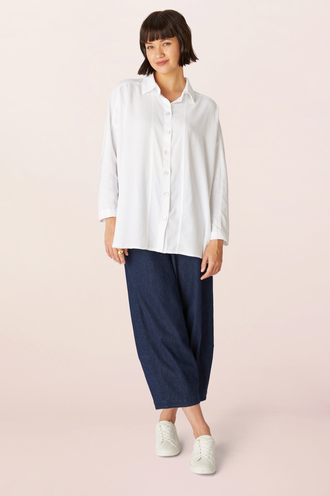 A woman stands against a light background wearing a Tencel Twill Boxy Shirt, wide-leg dark blue pants, and white sneakers.