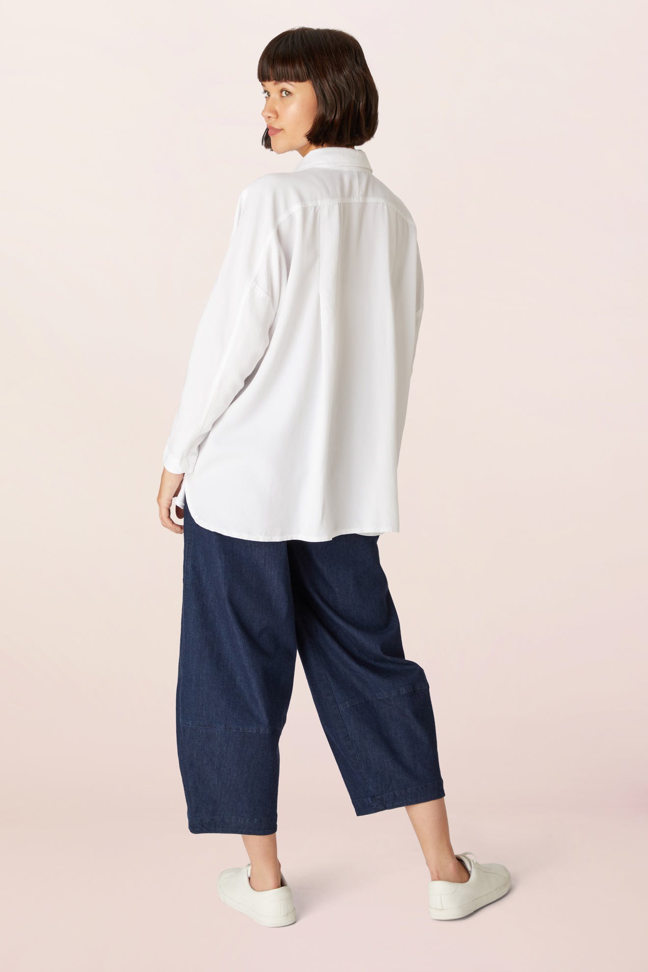 A person stands facing away, wearing a white Tencel Twill Boxy Shirt, wide-leg dark blue pants, and white sneakers against a light background.