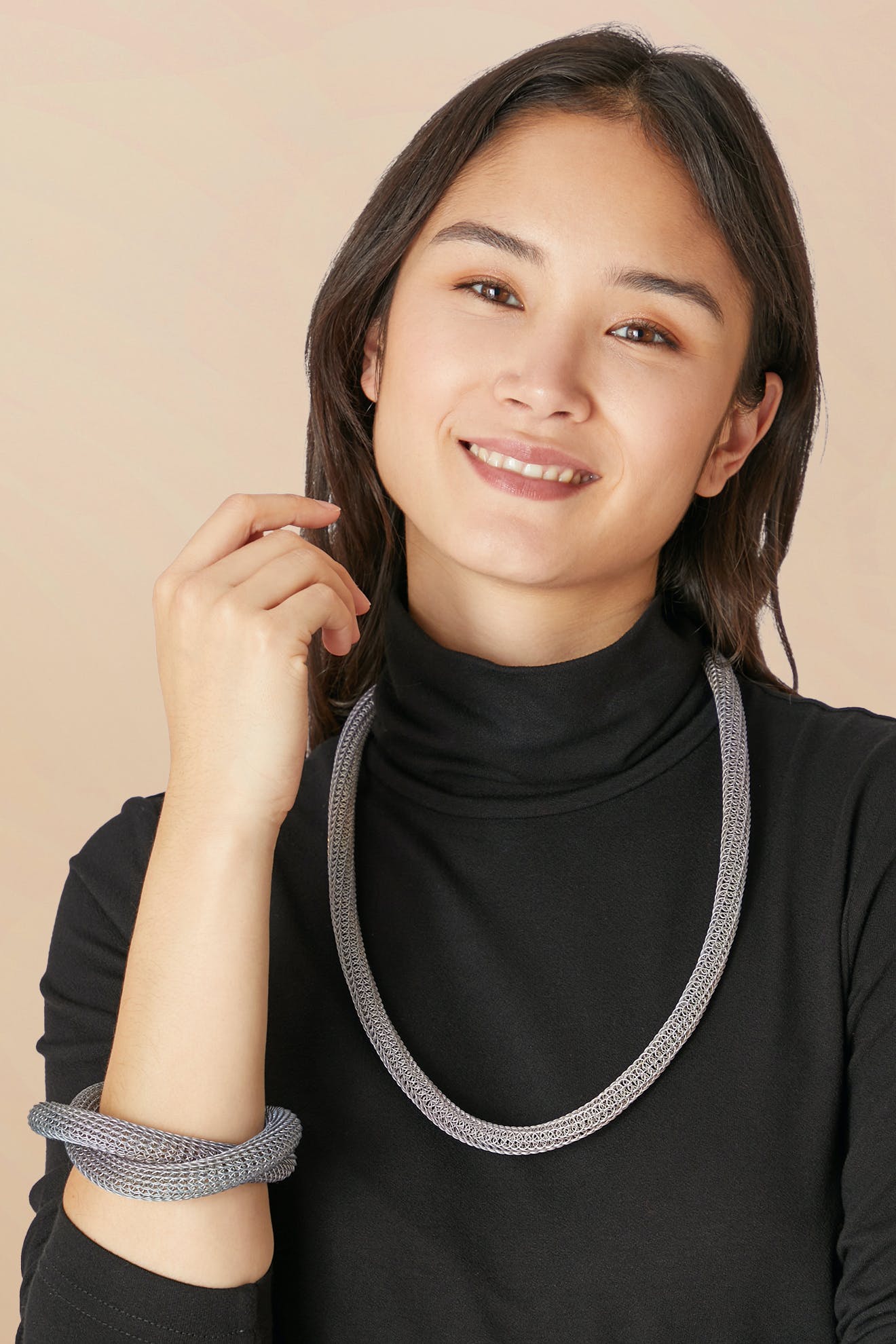 A woman smiles against a beige background, wearing a black turtleneck, silver jewelry, and the Knotted Bangle on her wrist, with one hand raised near her face.