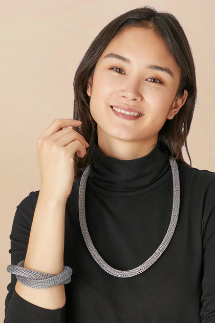 A woman smiles against a beige background, wearing a black turtleneck, silver jewelry, and the Knotted Bangle on her wrist, with one hand raised near her face.