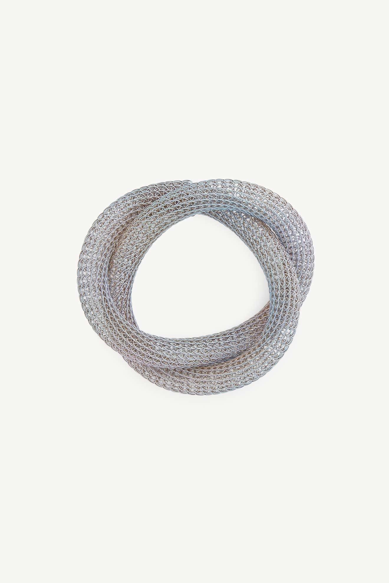 The Knotted Bangle, crafted in silver mesh, is coiled loosely against a plain white background, highlighting its elegant metalwork and refined design.