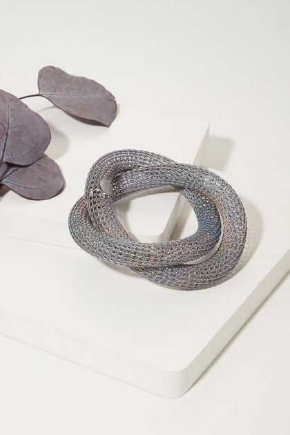 The Knotted Bangle, a handmade metallic mesh bracelet from the USA, is shown on a white surface beside dried purple leaves.