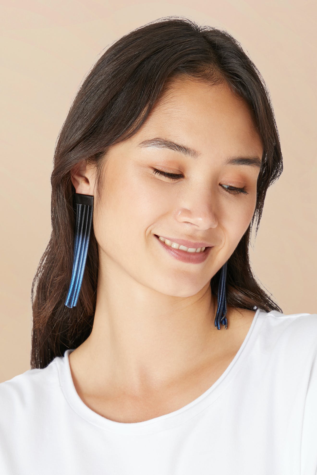 A woman with long dark hair wears a white shirt and Long Fringe Earrings, looking down and smiling slightly against a beige background.