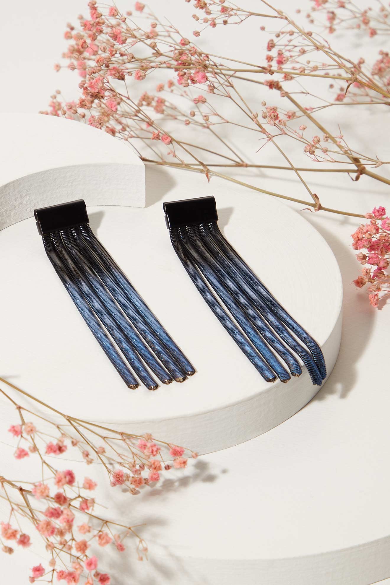 Long Fringe Earrings: a striking pair of blue mesh drop earrings with black tops, shown on a white circular platform and surrounded by pink dried flowers—unique metal jewellery that stands out.